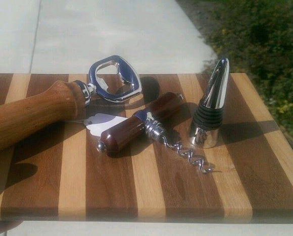 wooden bottle opener with metal head and T-handled bottle stopper, showing corkscrew on top of cutting board with alternating light and dark wood